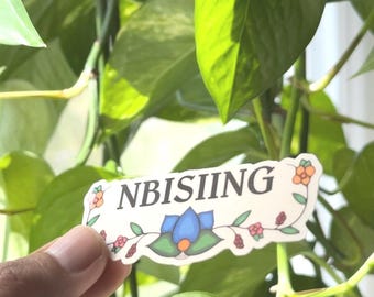 May include: A sticker with the word "NBISIING" in black letters, decorated with floral and leaf designs in orange, blue, and red. The sticker is held in front of a background of green leaves.