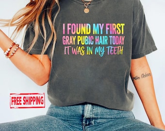 May include: A charcoal gray short-sleeve t-shirt featuring the multi-colored text I FOUND MY FIRST GRAY PUBIC HAIR TODAY IT WAS IN MY TEETH printed on the front. The shirt has a crew neckline and a relaxed fit, worn with blue denim jeans.