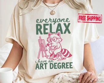 May include: A cream-coloured t-shirt featuring a graphic of a raccoon wearing glasses, sitting on a stool, and painting at an easel. The text reads "everyone RELAX i have an ART DEGREE" in dark green and pink lettering.