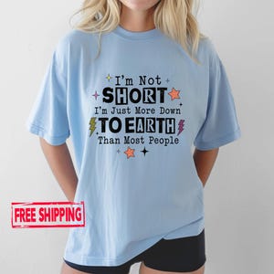 May include: A light blue short-sleeve t-shirt featuring the black text I'm Not SHORT I'm Just More Down TO EARTH Than Most People, accented with small orange and yellow star and lightning bolt graphics printed on the front of the garment.