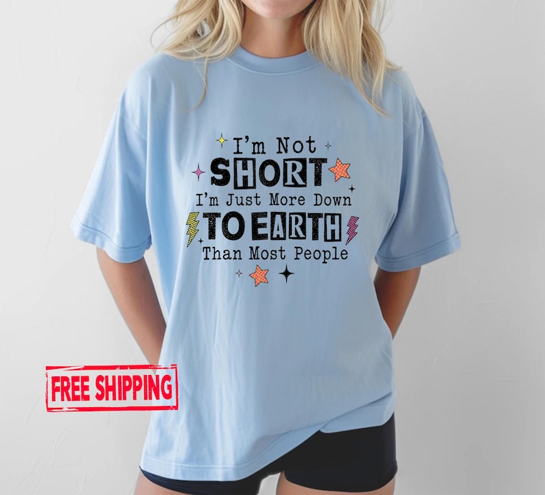 May include: A light blue short-sleeve t-shirt featuring the black text I'm Not SHORT I'm Just More Down TO EARTH Than Most People, accented with small orange and yellow star and lightning bolt graphics printed on the front of the garment.
