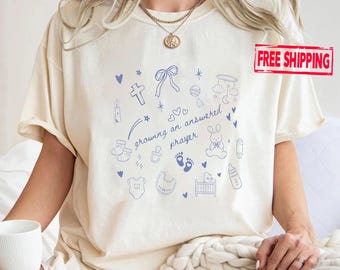 May include: A cream-coloured short-sleeve t-shirt featuring a blue graphic print of baby-themed icons including a cross, ribbon, rattle, mobile, pacifier, footprints, bunny, onesie, bib, crib, and bottle with the text growing an answered prayer.