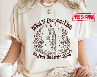 May include: A cream-coloured t-shirt featuring a dark brown graphic of a frog holding a cup, surrounded by mushrooms and stars. The text reads, What If Everyone Else Is Just Underthinking? The design is centred on the chest of the garment.