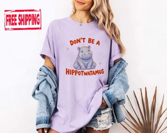 May include: A light purple short-sleeved t-shirt featuring a graphic of a blue hippopotamus in the center. The text above the hippo reads "DON'T BE A" and the text below reads "HIPPOTWATAMUS" in orange letters, accented by two small stars.