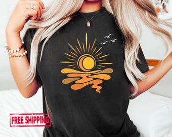 May include: A black short-sleeved t-shirt featuring a graphic print of a yellow sun with orange rays above stylised orange and yellow water waves. Three small white birds are flying to the right of the sun. The shirt has a crew neckline.
