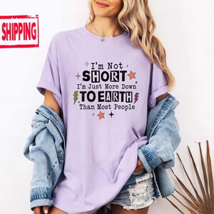 May include: A light purple t-shirt with black text that reads I'm Not SHORT I'm Just More Down TO EARTH Than Most People. The design includes small orange stars, yellow lightning bolts, and black star icons scattered around the printed lettering.