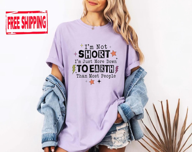 May include: A light purple t-shirt with black text that reads I'm Not SHORT I'm Just More Down TO EARTH Than Most People. The design includes small orange stars, yellow lightning bolts, and black star icons scattered around the printed lettering.