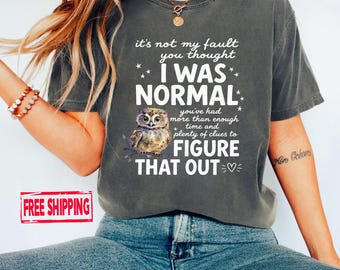 May include: A charcoal grey short-sleeve t-shirt featuring a graphic of a small owl and white text that reads: it's not my fault you thought I WAS NORMAL you've had more than enough time and plenty of clues to FIGURE THAT OUT with a heart icon.