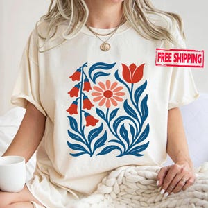 May include: Cream-colored t-shirt featuring a floral design with red bell-shaped flowers, a coral daisy, and a red tulip, all set against blue leaves and stems. The shirt has short sleeves and a relaxed fit.