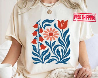 May include: A cream-coloured t-shirt with a floral design. The design includes red bell-shaped flowers, a coral daisy, and a red tulip, set against blue leaves and stems. The t-shirt has short sleeves and a relaxed fit.