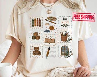 May include: Cream-colored t-shirt featuring a grid of illustrated elements including crayons, a paintbrush, a sun, an open book, and a teddy bear. The words "be kind" are also displayed. The shirt has a relaxed fit.