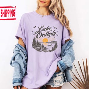 May include: A light purple t-shirt featuring a graphic print of Lake Ontario with a sunset, pine trees, and birds. The design includes the text Lake Ontario in a dark script font. The shirt is worn with a blue denim jacket and denim shorts.