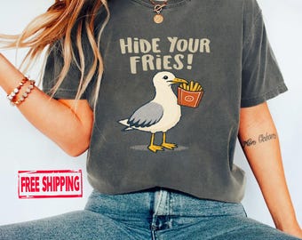 May include: A charcoal gray short-sleeve t-shirt featuring a graphic of a seagull holding a red carton of french fries. The text above the bird reads HIDE YOUR FRIES! in a light beige font. The shirt has a crew neckline and a relaxed fit.