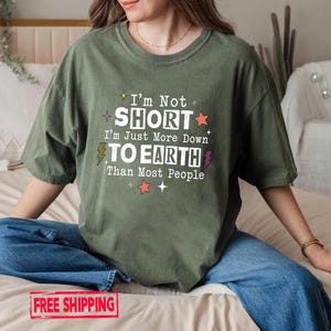 May include: An olive green short-sleeve t-shirt featuring white text that reads I'm Not SHORT I'm Just More Down TO EARTH Than Most People. The design includes small colorful star and lightning bolt graphics surrounding the printed text.