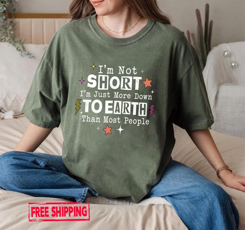 May include: An olive green short-sleeve t-shirt featuring white text that reads I'm Not SHORT I'm Just More Down TO EARTH Than Most People. The design includes small colorful star and lightning bolt graphics surrounding the printed text.