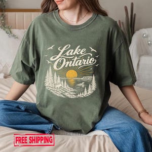 May include: An olive green t-shirt featuring a graphic print of a sunset over Lake Ontario. The design includes stylized pine trees, a moose, a bird on the water, and flying birds, with the text Lake Ontario written in a cursive script.