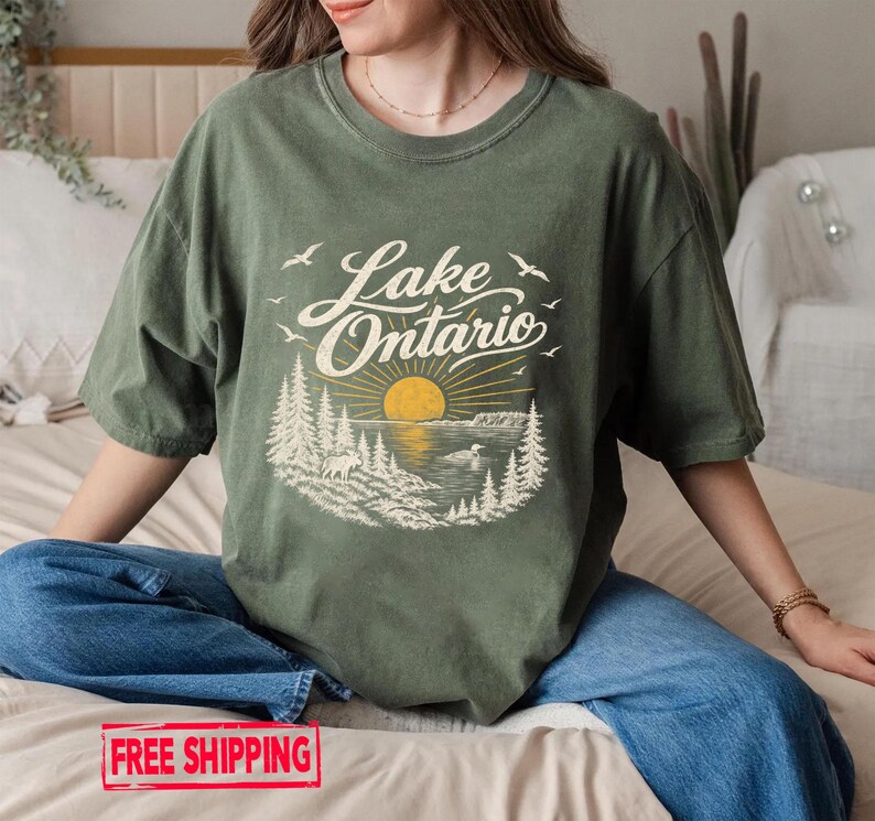 May include: An olive green t-shirt featuring a graphic print of a sunset over Lake Ontario. The design includes stylized pine trees, a moose, a bird on the water, and flying birds, with the text Lake Ontario written in a cursive script.