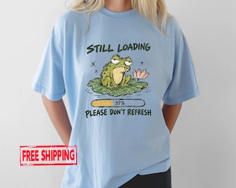 May include: A light blue t-shirt featuring a graphic of a green frog sitting on a lily pad. The text reads STILL LOADING above the frog and PLEASE DON'T REFRESH below a loading bar that shows 37%. Small dragonflies and a pink flower are visible.