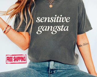 May include: Dark grey t-shirt with the words "sensitive gangsta" in white. The shirt is a crew neck and appears to be made of a soft material. The model is wearing blue jeans and a gold necklace.