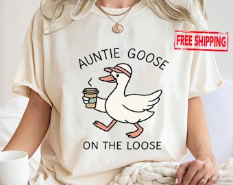 May include: Cream-colored t-shirt featuring a cartoon goose wearing a cap and holding a coffee cup. The text "AUNTIE GOOSE ON THE LOOSE" is printed above and below the goose illustration. A red rectangle with the words "FREE SHIPPING" is in the upper right corner.