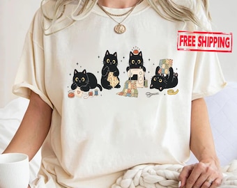 May include: A cream-colored t-shirt featuring a graphic of four black cats engaged in sewing activities. The cats are shown with sewing supplies including a sewing machine, fabric scraps, scissors, and thread spools. The design includes small star accents.