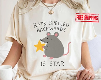 May include: Cream-colored t-shirt with the text "RATS SPELLED BACKWARDS IS STAR" above a cartoon illustration of a gray mouse holding a yellow star. The mouse has pink ears and a pink tail. The shirt is a casual style.