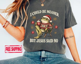 May include: A charcoal grey t-shirt featuring a graphic of a frog wearing a Santa hat and a red patterned jumper, sitting in a chair while holding a mug. The text reads I COULD BE MEANER BUT JESUS SAID NO, surrounded by snowflakes and ornaments.