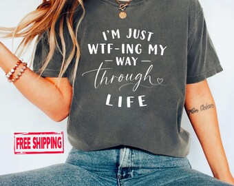 May include: A charcoal grey short-sleeved t-shirt featuring white text that reads I'M JUST WTF-ING MY WAY THROUGH LIFE with a small heart icon. The shirt has a crew neckline and a relaxed fit, showcasing a casual style with bold, centred lettering.