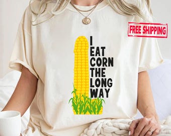 May include: Cream-colored t-shirt with the text "I EAT CORN THE LONG WAY" in black. A yellow corn graphic is centered on the shirt, with green grass at the bottom. The shirt has short sleeves.