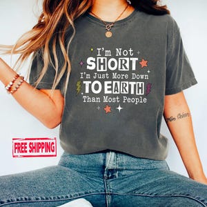 May include: A charcoal gray short-sleeve t-shirt featuring the white text I'm Not SHORT I'm Just More Down TO EARTH Than Most People, accented with small orange stars, yellow and purple lightning bolts, and white sparkles on the front.