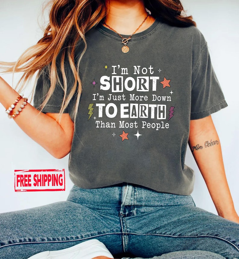May include: A charcoal gray short-sleeve t-shirt featuring the white text I'm Not SHORT I'm Just More Down TO EARTH Than Most People, accented with small orange stars, yellow and purple lightning bolts, and white sparkles on the front.