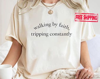 May include: A cream-coloured t-shirt featuring the text "walking by faith, tripping constantly" in black. The shirt has short sleeves and a loose fit. The t-shirt is being worn by a person.