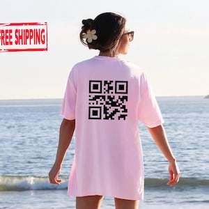 May include: A light pink t-shirt with a large black QR code printed on the back. The person is standing on a beach with the ocean in the background. The words "FREE SHIPPING" are in a red rectangle in the upper left corner.