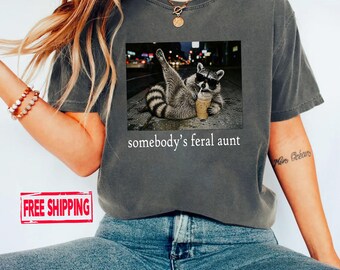 Könnte beinhalten: Ein anthrazitfarbenes Kurzarm-T-Shirt mit einem Grafikdruck eines Waschbären, der eine Sonnenbrille trägt und einen Plastikbecher mit Strohhalm hält. Unter dem Bild steht der Text somebody's feral aunt in einfacher weißer Schrift.