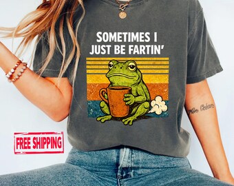 May include: Dark gray t-shirt featuring a cartoon frog holding a coffee mug, with the text "SOMETIMES I JUST BE FARTIN'". The frog is green, and the mug is brown. The background has horizontal stripes in orange, yellow, and blue.