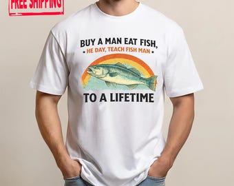 May include: White t-shirt with a colorful graphic of a fish jumping over a rainbow. The text reads "BUY A MAN EAT FISH, HE DAY, TEACH FISH MAN TO A LIFETIME". The shirt has a "FREE SHIPPING" label.