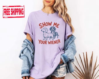 May include: A light purple crew neck t-shirt featuring a graphic of two dachshund dogs in dark blue ink. The text "SHOW ME YOUR WIENER" is printed in a bold, reddish-orange font above and below the dog illustration. The shirt has short sleeves.