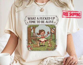 May include: Cream-coloured t-shirt with a framed illustration. The design shows a girl in a red and white dress in a forest scene with animals and the text "WHAT A FUCKED UP TIME TO BE ALIVE".