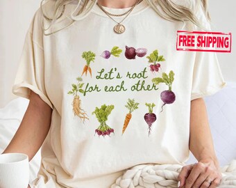 Könnte beinhalten: Ein cremefarbenes Kurzarm-T-Shirt mit einer kreisförmigen Anordnung verschiedener Wurzelgemüse wie Karotten, Radieschen und Rote Bete. Der Text "Let's root for each other" ist in grüner Schrift in der Mitte der Gemüseillustration aufgedruckt.
