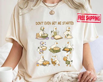 May include: A cream-colored t-shirt featuring a graphic print of nine white ducks in various baking activities. The text above the ducks reads, "DON'T EVEN GET ME STARTED." The ducks are shown mixing, kneading, and holding bread loaves.