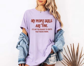 Könnte beinhalten: Ein hellviolettes T-Shirt mit dunkelrotem Text, der lautet: MY PEOPLE SKILLS ARE FINE. IT'S MY TOLERANCE TO IDIOTS THAT NEEDS WORK. Das T-Shirt hat kurze Ärmel und einen Rundhalsausschnitt, getragen mit einer blauen Jeansjacke und Jeansshorts.
