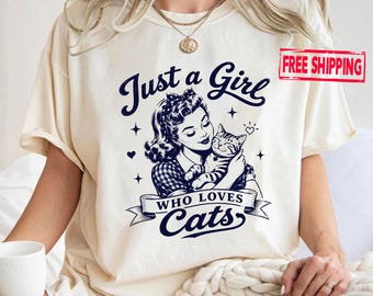 Könnte beinhalten: Ein cremefarbenes T-Shirt mit einem dunkelblauen Grafikdruck einer Person mit Vintage-Frisur, die eine Katze hält. Der Text lautet Just a Girl WHO LOVES Cats, ergänzt durch kleine Stern- und Herzsymbole. Das Motiv ist mittig platziert.