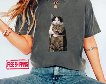 Könnte beinhalten: Ein anthrazitfarbenes Kurzarm-T-Shirt mit einem Grafikdruck einer schwarz-weißen Katze, die eine braune getigerte Katze umarmt. Die getigerte Katze hat den Mund offen und die Zunge ist sichtbar. Das Shirt hat einen Rundhalsausschnitt.