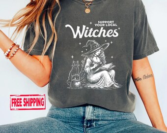Könnte beinhalten: Ein anthrazitfarbenes Kurzarm-T-Shirt mit einem weißen Grafikdruck einer Hexe mit spitzem Hut, die neben einer Katze und einem Kessel ein Buch liest. Der Text über der Illustration lautet in weißen Buchstaben SUPPORT YOUR LOCAL Witches.