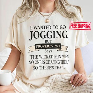 May include: Cream-colored t-shirt with the text "I WANTED TO GO JOGGING BUT PROVERBS 28:1 Says 'THE WICKED RUN WHEN NO ONE IS CHASING THEM.' SO THERE'S THAT..." printed in black. The shirt has short sleeves.