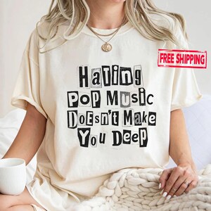 May include: A cream-colored short-sleeve t-shirt featuring black text in a ransom-note style that reads: Hating Pop Music Doesn't Make You Deep. The shirt has a crew neckline and is worn with a gold-toned chain necklace with a circular pendant.