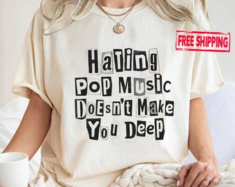 May include: A cream-coloured short-sleeve t-shirt featuring black text in a ransom-note style that reads: Hating Pop Music Doesn't Make You Deep. The shirt has a crew neckline and is worn with a gold-toned chain necklace with a circular pendant.