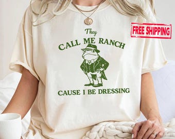 May include: A cream-coloured short-sleeve t-shirt featuring a dark green graphic of a frog wearing a suit, hat, and bowtie. The text above the frog reads "They CALL ME RANCH" and the text below reads "CAUSE I BE DRESSING" in a dark green font.