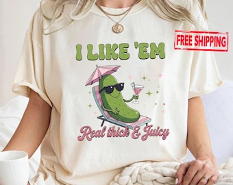 May include: A cream-colored t-shirt featuring a graphic of a green pickle wearing black sunglasses, lounging on a beach chair under a pink umbrella while holding a cocktail. The text reads I LIKE 'EM and Real thick & Juicy in green and pink.