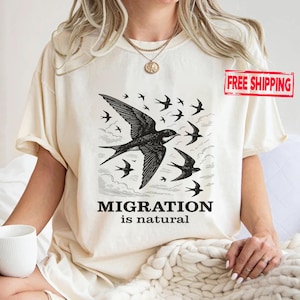 May include: Cream-colored t-shirt featuring a black illustration of swallows in flight. The design includes the text "MIGRATION is natural" in a bold, sans-serif font. The shirt has short sleeves and a relaxed fit.